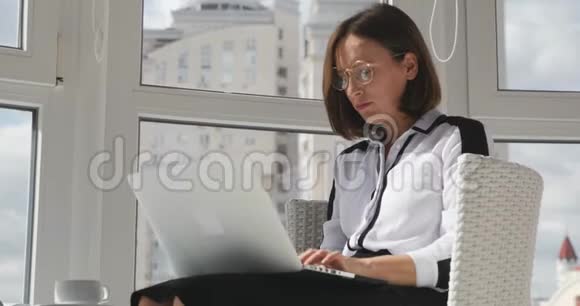 自信的女性穿着白色衬衫和眼镜在现代办公室工作在笔记本电脑上工作有大窗户打字电子邮件集中视频的预览图