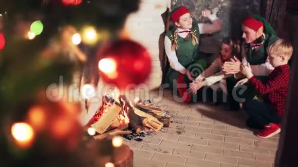 快乐的孩子们和圣诞老人的精灵们在壁炉旁温暖他们的手视频的预览图