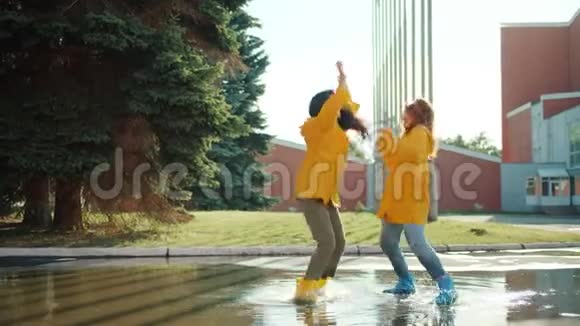 快乐的女孩朋友们穿着雨衣和雨靴在水坑里跳着舞视频的预览图