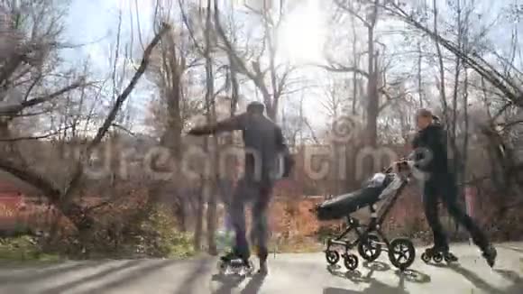 年轻幸福的家庭享受在温暖的轮滑公园散步与幼儿在婴儿车爸爸倒着骑是的视频的预览图