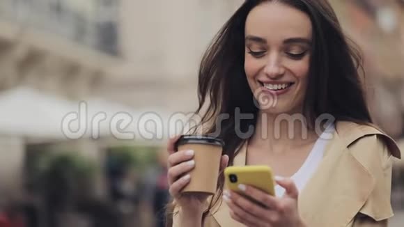 年轻漂亮的女孩打字时她的智能手机和微笑手里拿着一个纸杯喝咖啡站在老城区视频的预览图