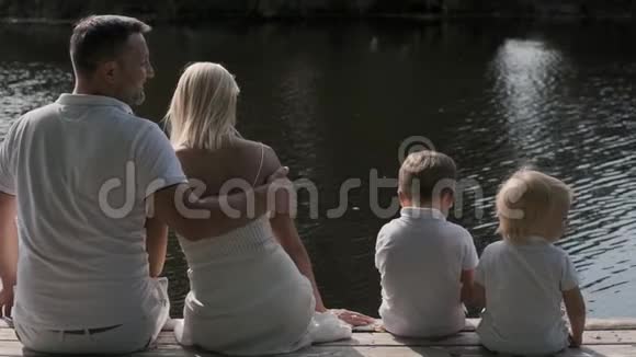 幸福的家庭爸爸妈妈和两个孩子坐在河边的码头上背面视频的预览图