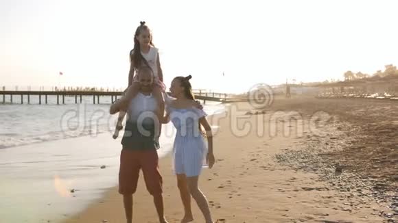 快乐的父亲和她的两个女儿在日落时在海滩上玩得很开心有个女儿坐在他肩膀上的男人视频的预览图