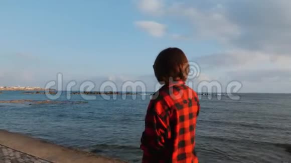 日落时分身着红色格子衬衫的年轻黑发女子沿着旅游长廊散步美丽的女孩在海边享受夜晚视频的预览图