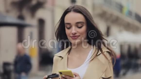 年轻漂亮的女孩拿着咖啡杯微笑使用她的智能手机和打字走在城市街道靠近右侧视频的预览图