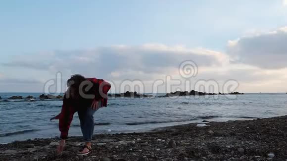 忧郁的年轻女性穿着温暖的红色衬衫和牛仔裤在岩石海滩上收集石头把它们扔进黑暗的大海视频的预览图