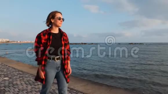 晚上穿着红色格子衬衫和牛仔裤沿着海滨散步的可爱的黑发女性年轻女孩走在空荡荡的长廊上视频的预览图
