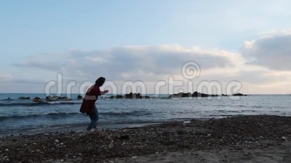 可爱的孤独的女人穿着牛仔裤和温暖的红色衬衫站在空荡荡的岩石海滩上在日落时把石头扔进水里视频的预览图
