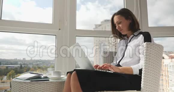 聪明成功的年轻女商人穿着白色衬衫和黑色裙子在有大窗户的工作场所工作笔记本电脑视频的预览图