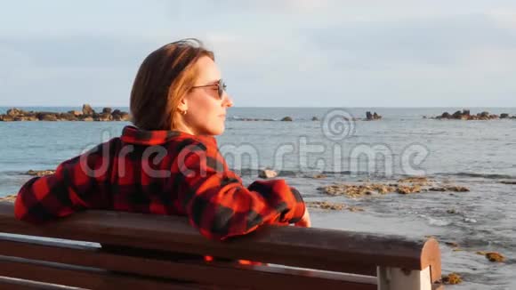 孤独忧郁的黑发女人坐在空荡荡的长廊的木凳上看着大海和日落靠近后景视频的预览图