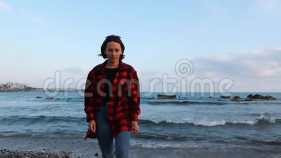 穿着红色温暖格子衬衫和牛仔裤的白种人浅黑肤色的年轻女性在风和日丽的夜晚漫步在空旷的海滩上视频的预览图