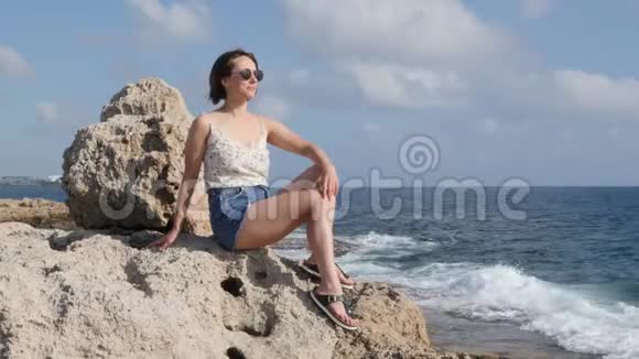 穿着牛仔裤短裤和黑色太阳镜坐在海边的岩石上看着地平线的性感女性视频的预览图