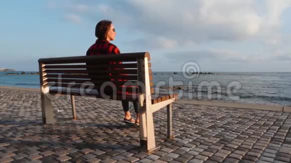 孤独悲伤的女性穿着温暖的格子衬衫和牛仔裤坐在空荡荡的码头上的木凳上看着夕阳视频的预览图