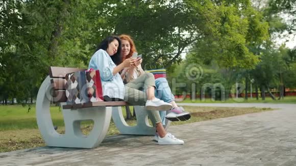 漂亮的女孩十几岁的青少年在公园的长椅上玩智能手机视频的预览图