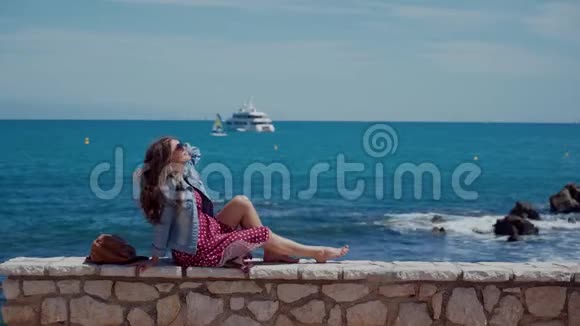 阳光明媚的日子里一位戴着墨镜穿着红色连衣裙的快乐年轻女子坐在石篱笆墙上那里有着迷人的大海和视频的预览图
