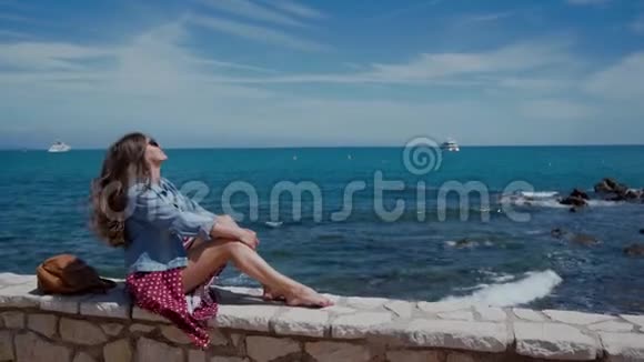 阳光明媚的一天穿着连衣裙和牛仔夹克的年轻女子坐在石栅栏上看着蔚蓝的大海风的美丽发展视频的预览图