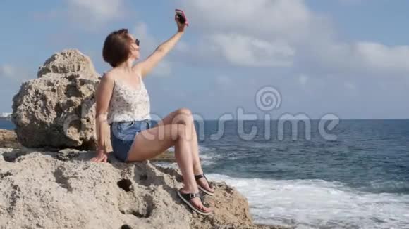 穿牛仔裤短裤的性感黑发女人在岩石海岸做自拍智能手机很有魅力的女性在自己身上拍照视频的预览图