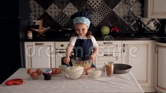 一个穿着围裙和帽子的小女孩在家里厨房里学做面团视频的预览图