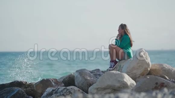 穿着运动鞋和绿色运动夹克的年轻女孩若有所思地坐在岩石沙滩上浪花溅在岩石上视频的预览图