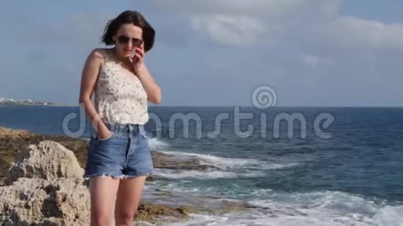 布鲁内特小姐穿着牛仔裤短裤和丝绸上衣站在岩石海岸的边缘打电话微笑着视频的预览图