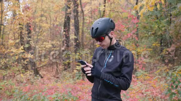 在秋天的森林里骑自行车训练前戴着黑色头盔和戴着运动眼镜的男性自行车运动员在电话中输入信息视频的预览图