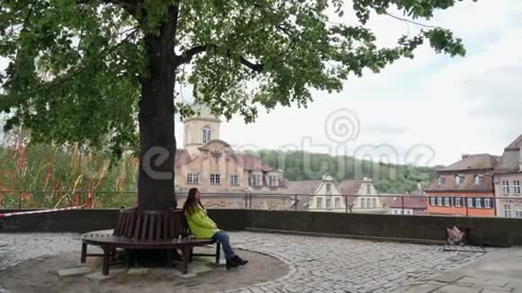 穿着黄色外套的漂亮女孩独自坐在公园或花园的长凳上坐在一棵高大的大树下俯瞰五颜六色的景色视频的预览图