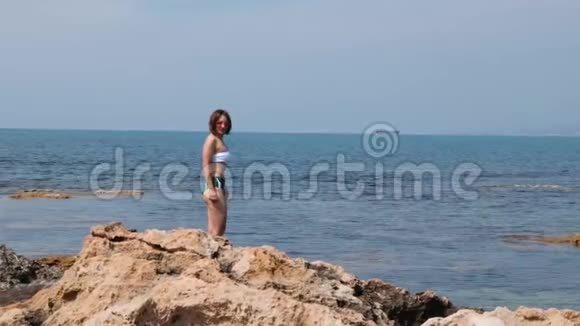性感迷人的黑发女人穿着比基尼和粉红色太阳镜站在海边的岩石海岸年轻女性的肖像视频的预览图