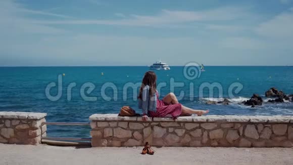 美丽的女人坐在法国南部安蒂布斯镇上方的石栅栏上欣赏美丽的海景遥远的计划视频的预览图