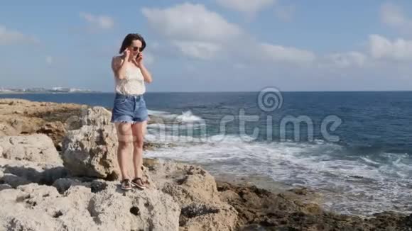 可爱的白种人年轻的黑发女人穿着牛仔裤短裤上衣站在海滨的边缘打电话视频的预览图