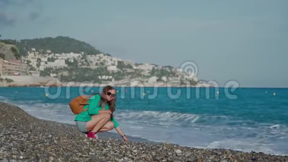 阳光明媚的一天穿着绿色夹克牛仔短裤和运动鞋的年轻快乐的女士背着背包坐在海边视频的预览图