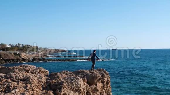 漂亮的白种人穿着蓝色衬衫和牛仔裤站在海边的悬崖边站在岩石上的年轻人视频的预览图