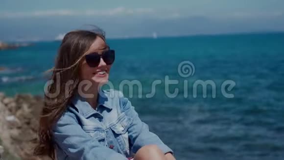 戴墨镜和牛仔夹克的微笑的女人坐在度假城市安蒂布的海边直发视频的预览图