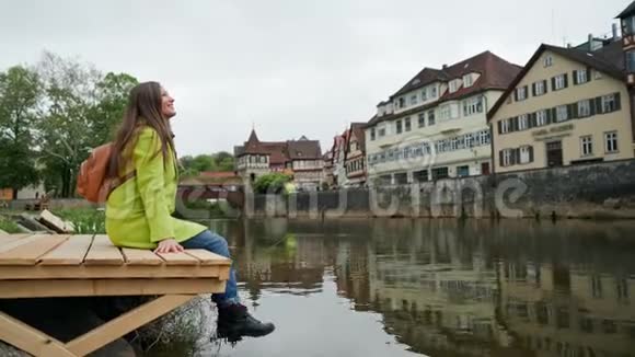穿着黄色外套的女孩背着背包坐在靠近水的木墩上看着传统的德国式半身视频的预览图