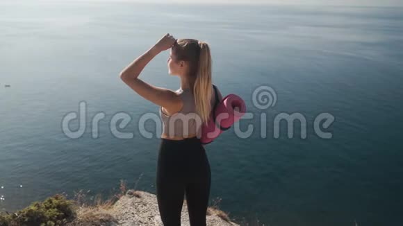 女人拿着瑜伽垫从悬崖上看海深呼吸慢动作视频的预览图