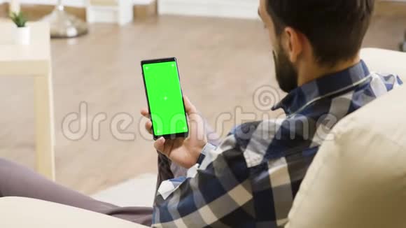 年轻人坐在沙发上看着上面有绿色屏幕的智能手机视频的预览图