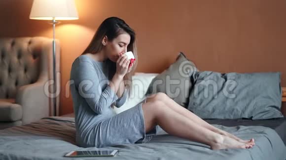快乐可爱的休闲女士穿着礼服喝着芬芳的热饮料坐在沙发上视频的预览图
