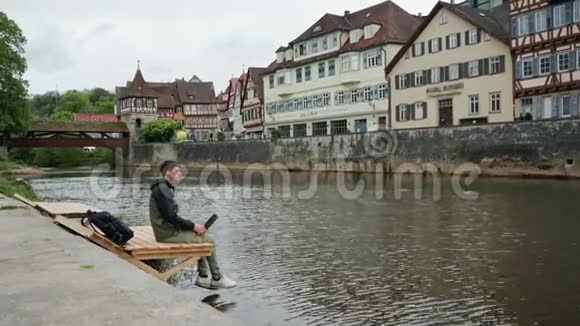 带背包的年轻人坐在靠近水的木墩上用笔记本传统德语视频的预览图