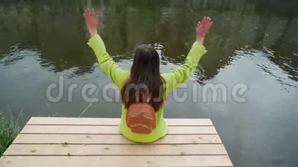 穿着黄色夹克的长发女人背包坐在河边或池塘的木墩上挥舞着双手传统视频的预览图