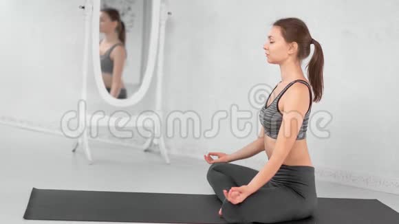 瑜伽女人坐在莲花的位置在白色房间的垫子上放松视频的预览图