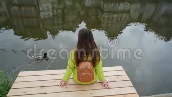 长头发的女人穿着黄色夹克背着背包坐在河边或池塘的木墩上看上去像鸭子在游泳传统视频的预览图