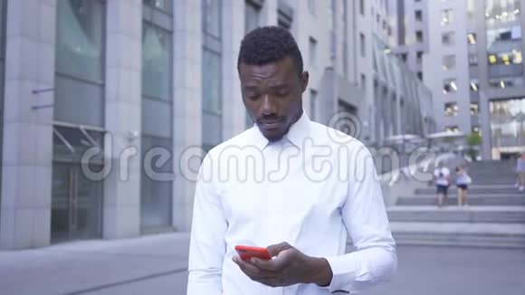 那个穿着白色衬衫的美国黑人男子站在城市街道上用红色手机说话男性商人或视频的预览图