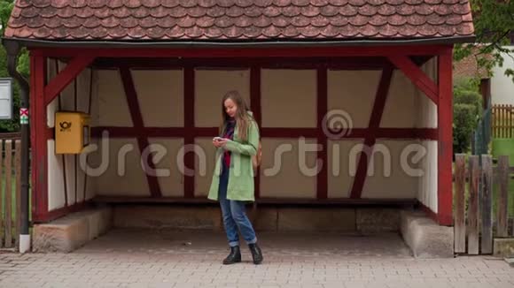 一位穿着绿色外套和背包的漂亮女士在Fachwerk风格的汽车站附近等公共汽车时使用智能手机视频的预览图