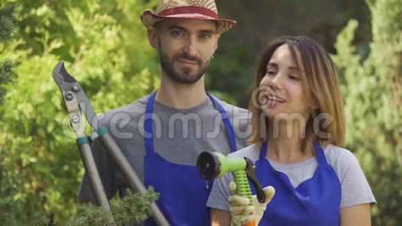 肖像微笑的白种人女人和穿着蓝色制服的英俊男子站在花园里拿着花园工具看着视频的预览图