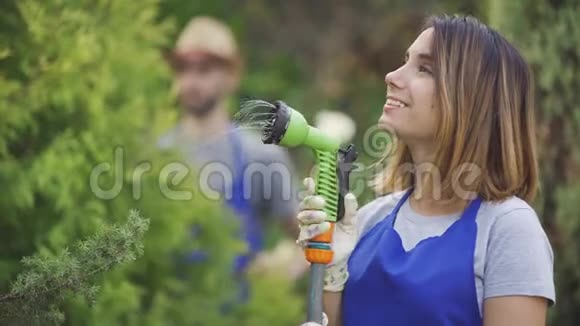 一幅美丽的高加索女人在前景中玩弄水管而男人在砍树叶时的画像视频的预览图