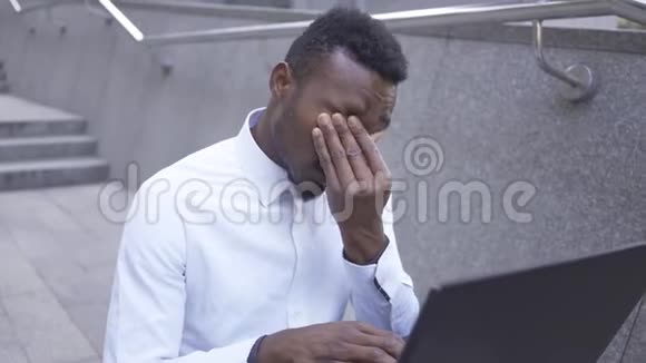 精疲力竭的非裔美国人穿着白色衬衫揉着眼睛在笔记本电脑上打字厌倦了男性商人或经理视频的预览图