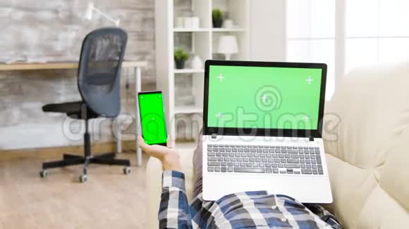视差镜头躺在沙发上手里拿着一部绿色屏幕智能手机和笔记本电脑视频的预览图