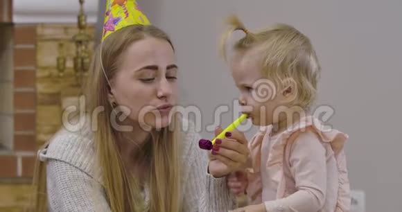 一幅美丽的白种人女人和小可爱女孩在家玩派对哨子的特写肖像年轻的母亲视频的预览图