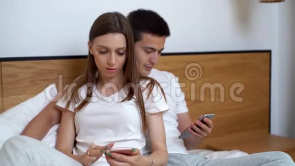 年轻的幸福夫妇谈笑风生着智能手机用有趣的应用程序坐在床上视频的预览图