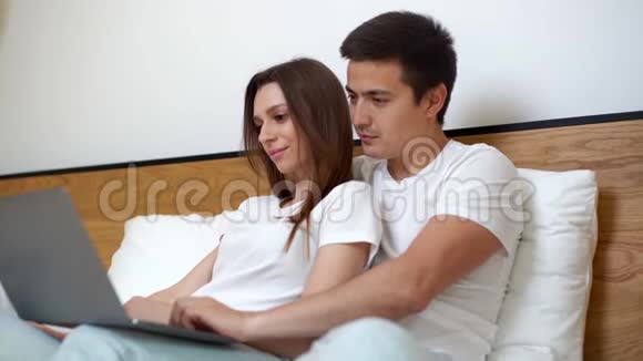 快乐的年轻夫妇躺在床上用笔记本电脑看电影视频的预览图