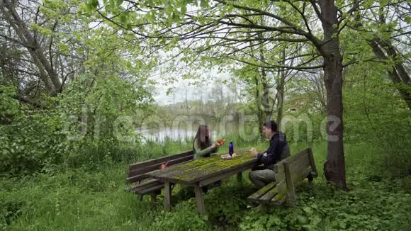 快乐的女孩和男人坐在湖边绿色公园的苔藓覆盖的野餐长凳上穿过布瑞泽尔的女孩视频的预览图